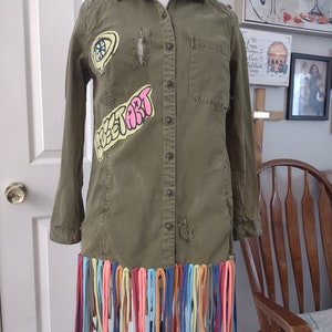May include: An olive green button-down shirt dress with patches that read "Sweet Art" and a design. The bottom hem is adorned with colorful fabric fringe. The dress is displayed on a mannequin.