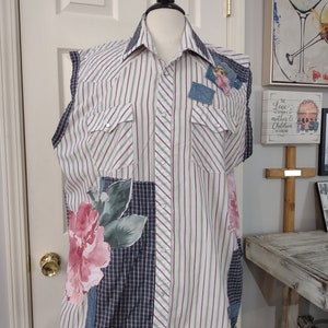 May include: A sleeveless shirt on a mannequin. The shirt is a patchwork design with vertical stripes, plaid, and floral patterns. It features a large pink flower and denim accents. The shirt has two chest pockets and silver snap closures.