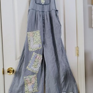 May include: Pale blue denim dungarees with wide legs, hanging on a hanger. The overalls feature floral and patchwork details, including a flower on the bib and patches spelling "LOVE" on the legs, plus a heart patch.