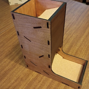May include: A wooden dice tower with a natural wood finish. The tower has a tall, rectangular body with a tray at the bottom. The tower is constructed with interlocking pieces and features small, dark accents. The interior of the tower and tray are lined with a light-colored material.