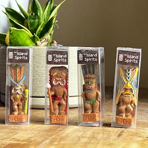 Könnte beinhalten: Vier kleine, farbenfrohe Tiki-Statuen in durchsichtiger Kunststoffverpackung. Jede Statue ist braun mit einzigartigen bemalten Details, darunter blaue, gelbe und rote Akzente. Die Verpackung trägt die Aufschrift "Island Spirits". Eine Topfpflanze befindet sich im Hintergrund.
