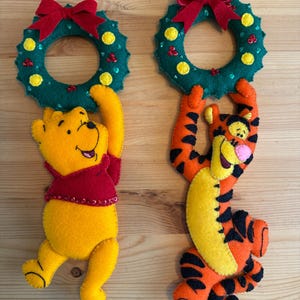 Puede incluir: Dos adornos navideños de fieltro con Winnie the Pooh y Tigger. Cada personaje sostiene una corona verde con un lazo rojo. Los adornos son amarillos y naranjas, con detalles rojos y negros. Las coronas tienen adornos amarillos y rojos.
