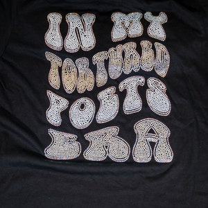 May include: Black t-shirt with the phrase "IN MY FORTIES I'M NOT TOO OLD FOR THIS SH*T" spelled out in a bubbly, rhinestone-studded font. The text is the focal point, creating a bold statement on the dark fabric.