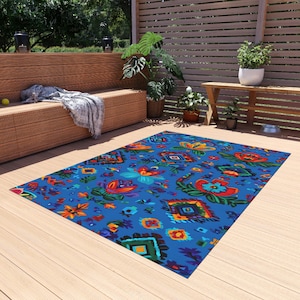 Puede incluir: Una alfombra rectangular azul para exteriores con un vibrante estampado floral. La alfombra presenta flores rojas, naranjas y amarillas, junto con formas geométricas en varios colores. La alfombra está colocada sobre una terraza de madera.