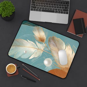 May include: A teal mouse mat with a design of gold and white feathers and pearl accents. A white computer mouse sits on the mat, with a laptop and other stationery visible in the background.