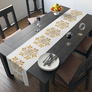 May include: A white table runner featuring a repeating pattern of gold and grey floral snowflake-like motifs. The runner is laid out on a dark wooden dining table set with white plates, silverware, wine glasses, and a bowl of apples.