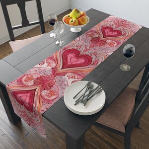 Op de afbeelding: Een decoratieve tafelloper met een hartvormig ontwerp in roze en rode tinten. De loper ligt op een donkere houten tafel, samen met een wit bord, bestek en wijnglazen. Een kom met appels is zichtbaar op de achtergrond.