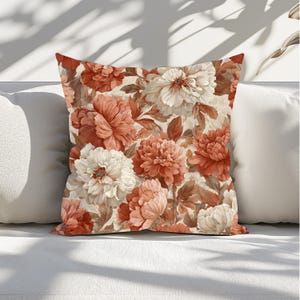 May include: Square decorative pillow with a floral pattern. The pillow features large, stylized flowers in shades of coral, cream, and brown against a cream background. The pillow is resting on a white surface.