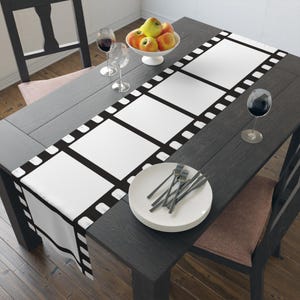 May include: A black dining table is set with a white film strip-patterned table runner. The runner has large white rectangles bordered by black, with white rounded edges. A white plate with silverware, wine glasses, and a bowl of fruit complete the setting.