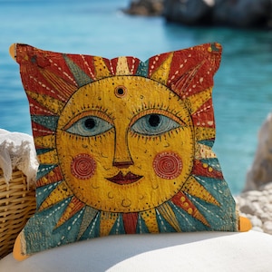 May include: A square throw pillow featuring a vibrant yellow sun illustration with large blue eyes, red spiral cheeks, and a third eye on the forehead. The sun is surrounded by alternating red, yellow, and blue triangular rays on a textured fabric.