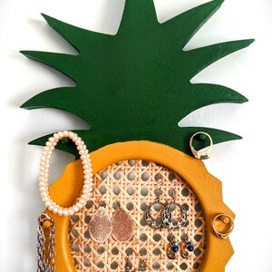 May include: A wooden pineapple-shaped jewelry organizer. The top is green, and the base is yellow with a woven rattan center. Various earrings, rings, and a pearl bracelet hang from the organizer. A stylish way to display jewelry.