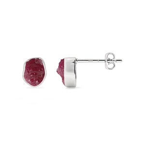 May include: A pair of stud earrings featuring raw, deep red ruby gemstones set in polished silver. The rubies have a natural, irregular shape, and the silver settings have a smooth, modern design. The earrings are displayed against a white background.