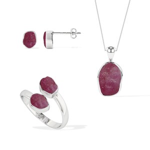 May include: A jewelry set featuring a necklace, ring, and earrings. The set includes a silver necklace with a pendant, a silver ring, and silver earrings. Each piece features a raw, deep red gemstone.