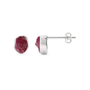 May include: A pair of stud earrings featuring raw, deep pink ruby stones set in polished silver. One earring is shown from the front, while the other is displayed from the side, revealing the silver post and butterfly back closure stamped with 925.
