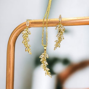 以下が含まれることがあります： 繊細な葉のデザインと透明なキュービックジルコニアの石が施された、ゴールドのネックレスとイヤリングのセット。ジュエリーは銅色のスタンドに飾られています。