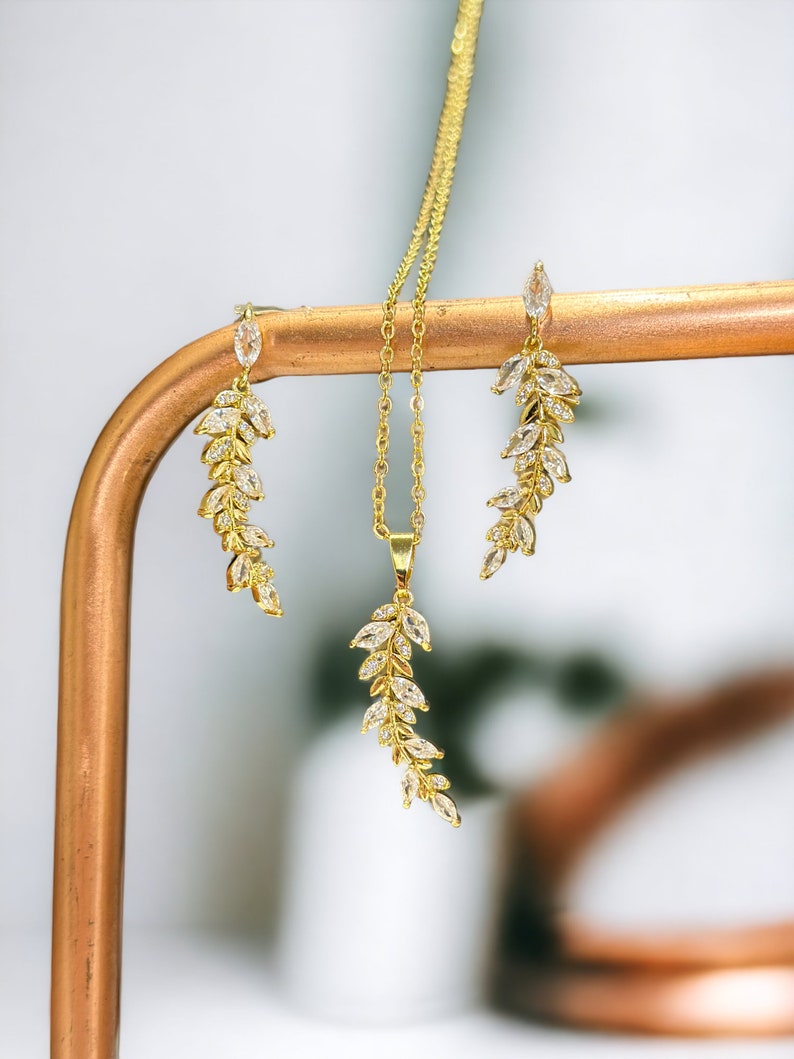 Pu&ograve; includere: Un set di collana e orecchini in oro con un delicato design a foglia e pietre di zircone cubico trasparenti. I gioielli sono esposti su un supporto color rame.