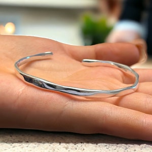 Może przedstawiać: Srebrna bransoletka typu bangle z młotkowanym, fakturowanym wykończeniem. Bransoletka jest otwarta i ma lekko zakrzywiony kształt.