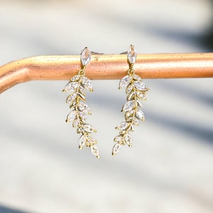 Pu&ograve; includere: Un paio di orecchini dorati con un delicato design a foglia. Ogni orecchino presenta una cascata di cristalli trasparenti a forma di marquise.