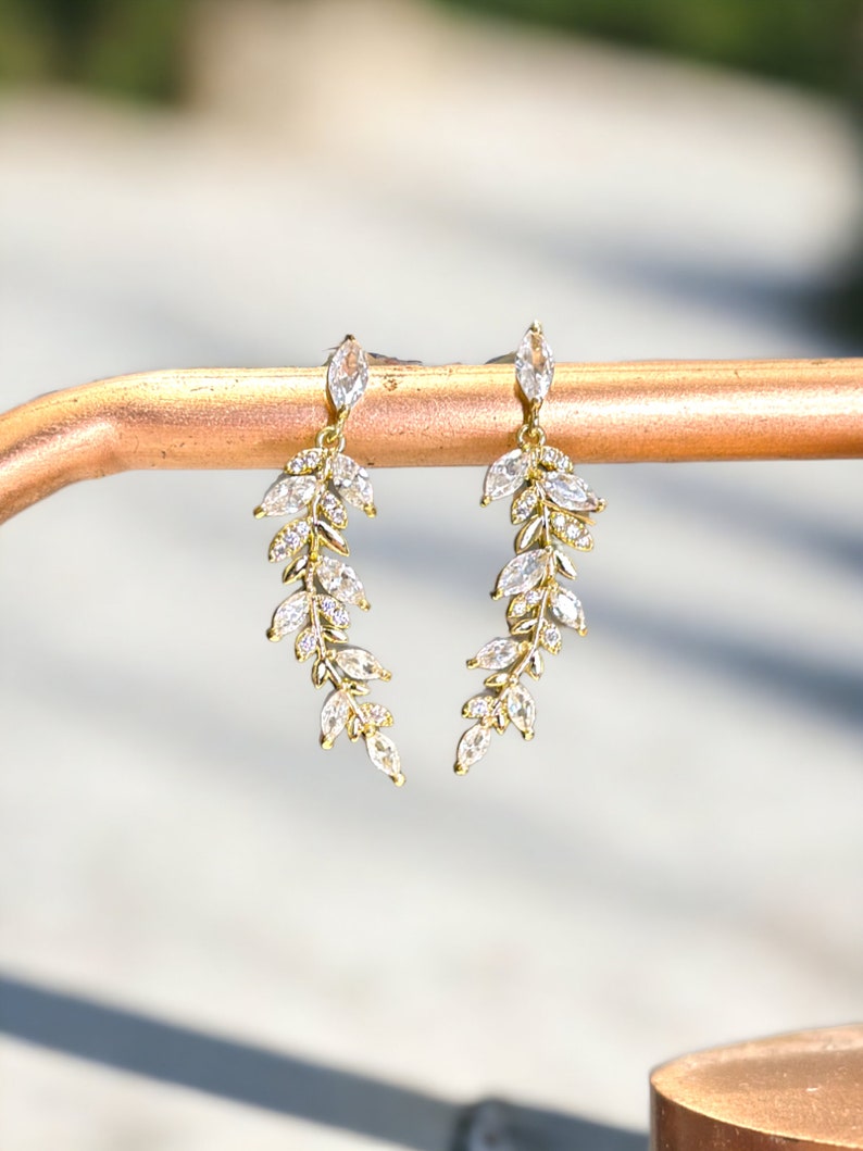 Pu&ograve; includere: Un paio di orecchini dorati con un delicato design a foglia. Ogni orecchino presenta una cascata di cristalli trasparenti a forma di marquise.