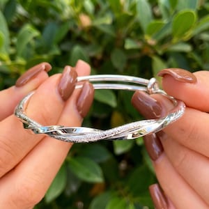 May include: A silver-toned expandable bangle bracelet featuring a twisted design. The bracelet consists of two intertwined strands, one with a smooth, polished finish and the other with a textured, sparkling surface, creating a layered appearance.
