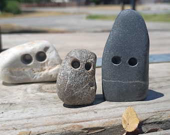 May include: Three smooth, rounded stones of varying sizes and colors, each featuring two small, circular, drilled holes that resemble eyes. The stones are light gray, speckled brown, and dark charcoal gray, arranged in a horizontal line.