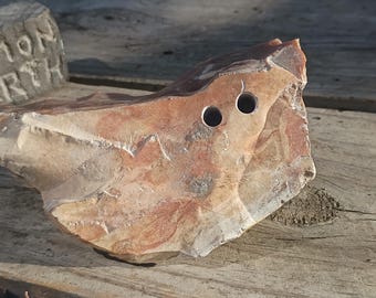 May include: A jagged, multi-colored stone fragment featuring shades of tan, brown, and reddish-orange with two circular holes drilled through the upper section. A separate gray stone block with the carved text SIMON EARTH is visible nearby.