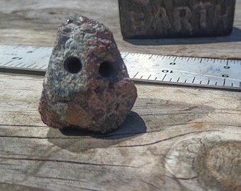 May include: A small, textured, multi-colored stone with two circular holes drilled into its front surface. The stone is positioned next to a metal ruler showing a measurement of approximately 1.5 inches in width at its widest point.