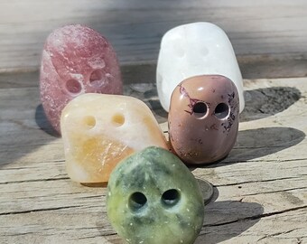 May include: A collection of five smooth, polished stones, each featuring two drilled holes. The stones vary in color, including shades of pink, white, yellow, green, and a mottled brown, arranged in a cluster on a weathered wooden surface.