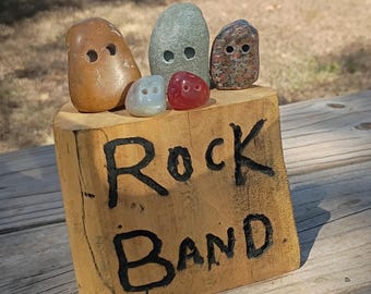 May include: A light brown wooden block with the text "Rock BAND." burned into the front. Five smooth, rounded stones are arranged on top of the block. Each stone features two small, circular holes drilled into the front surface.