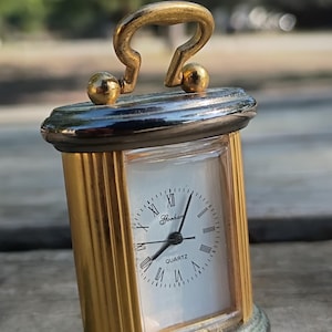 May include: A small, rectangular quartz clock with a gold-colored frame and a white face. The clock has black Roman numerals and hands, with a gold handle at the top. The clock is approximately 3 inches tall.