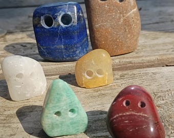 May include: A collection of six polished stones in various shapes and colors, including blue, brown, white, yellow, green, and red. Each stone features two small, circular holes drilled into the front surface, resembling a pair of eyes.