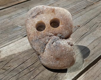 May include: A smooth, tan-colored river stone with a rounded, irregular shape. The surface of the stone features two distinct, circular, drilled holes positioned near the top, with natural dark brown mineral veins and textured weathering throughout.