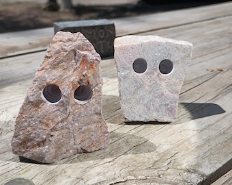 May include: Two rough-hewn stone sculptures with natural textures and colors. The left stone is reddish-brown with jagged edges, while the right stone is light grey and flatter. Both feature two circular, dark, hollowed-out holes drilled into them.