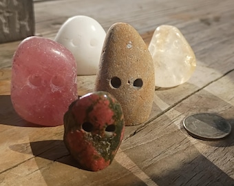 May include: A collection of five smooth, rounded stones, each featuring two small drilled holes. The stones vary in color, including pink, white, tan, translucent yellow, and a mottled red and green stone, displayed alongside a silver coin.