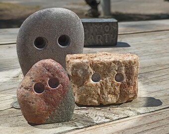 May include: Three natural stones with drilled holes, including a large smooth gray oval stone, a smaller reddish-brown stone, and a rectangular tan textured stone. A dark gray block with the engraved text SIMON EARTH is visible in the background.