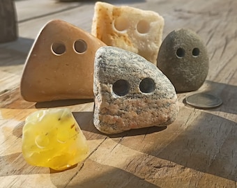 May include: A collection of five natural stones shaped into buttons, each featuring two drilled holes. The set includes a smooth tan triangular stone, a textured white stone, a speckled grey stone, a dark grey oval stone, and a translucent yellow stone.