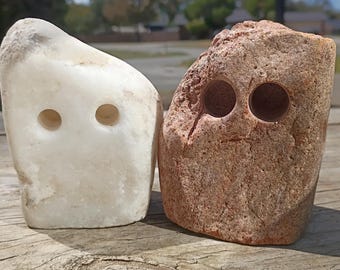 May include: Two irregular, roughly rectangular stones standing side by side. The stone on the left is smooth, off-white, and features two circular, drilled holes. The stone on the right is textured, reddish-brown, and also has two circular, drilled holes.
