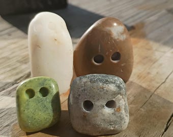 May include: Four smooth, rounded stones shaped like buttons, each featuring two drilled holes. The collection includes one tall white stone, one medium brown stone, one small speckled green stone, and one medium gray stone with a textured surface.