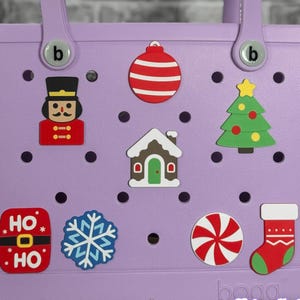 May include: A light purple tote bag decorated with Christmas-themed charms. Charms include a nutcracker, ornament, house, Christmas tree, snowflake, candy cane, stocking, and a "HO HO" design. The bag has a handle and the brand name "bogg" is visible.