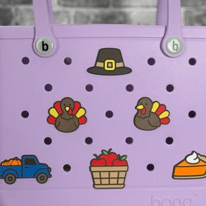 May include: A light purple tote bag with two handles and a variety of Thanksgiving-themed charms. The charms include a pilgrim hat, turkeys, a truck with pumpkins, a basket of apples, and a slice of pie.