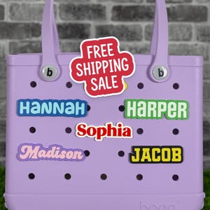 May include: A light purple tote bag with a handle and the brand name "bogg" on the bottom. The bag has several name patches with names like "Hannah", "Harper", "Sophia", "Madison", and "Jacob". A red and white "FREE SHIPPING SALE" sticker is also on the bag.