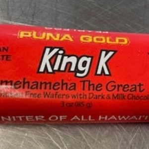 May include: A red chocolate bar wrapper with the text "PUNA GOLD" and "King K Kamehameha The Great" in white. The wrapper also features a drawing of a Hawaiian king in a red and yellow robe. The text "Layers of Ulu Gluten Free Wafers with Dark & Milk Chocolate" and "3 oz (85 g)" is printed on the wrapper. The text "UNITER OF ALL HAWAII'" is printed at the bottom of the wrapper.