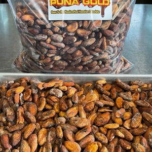 May include: A clear plastic bag filled with brown cocoa beans, labeled "PUNA GOLD Bean-To-Bar Handcrafted Hawaiian Chocolate." Below, a metal tray holds a pile of similar cocoa beans, showcasing their natural color and texture. The image highlights the raw ingredients for chocolate making.