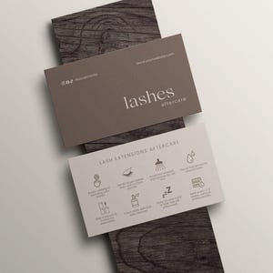 以下が含まれることがあります： 2つの名刺。 上部のカードは茶色で、「lashes aftercare」と書かれています。 下部のカードは白で、まつげエクステのアフターケアの手順とアイコンが記載されています。 カードは木製の表面に置かれています。