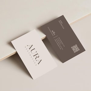 以下が含まれることがあります： AURA AESTHETICSという文字が記された2枚の長方形の名刺。1枚はオフホワイト地に濃い文字、もう1枚はダークブラウン地に白い文字で、連絡先情報、ソーシャルメディアアイコン、正方形のQRコードが記載されています。
