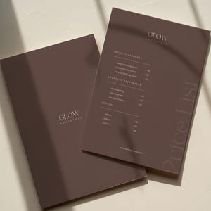 Könnte beinhalten: Zwei braune Broschüren mit dem Text "GLOW AESTHETIC" und "Price List". Die Broschüre rechts zeigt eine Liste von Gesichts- und erweiterten Behandlungen. Die Broschüren liegen auf einer hellbeige Oberfläche mit Schatten.