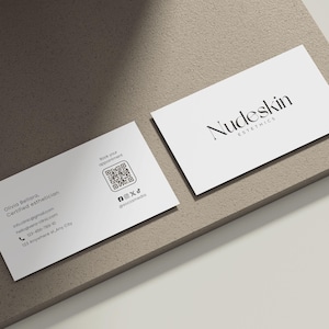 May include: Two white business cards with the text "Nudeskin Esthetics" and contact information. One card displays the business name, while the other includes a QR code and social media handles. The cards are on a neutral-toned surface.