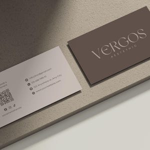 以下が含まれることがあります： 2 つの名刺。1 つは「VERGOS AESTHETIC」の白文字が入ったトープ色、もう 1 つは濃い灰色の文字と連絡先情報が入った薄いピンク色です。カードは、質感のある明るい茶色の表面にあります。
