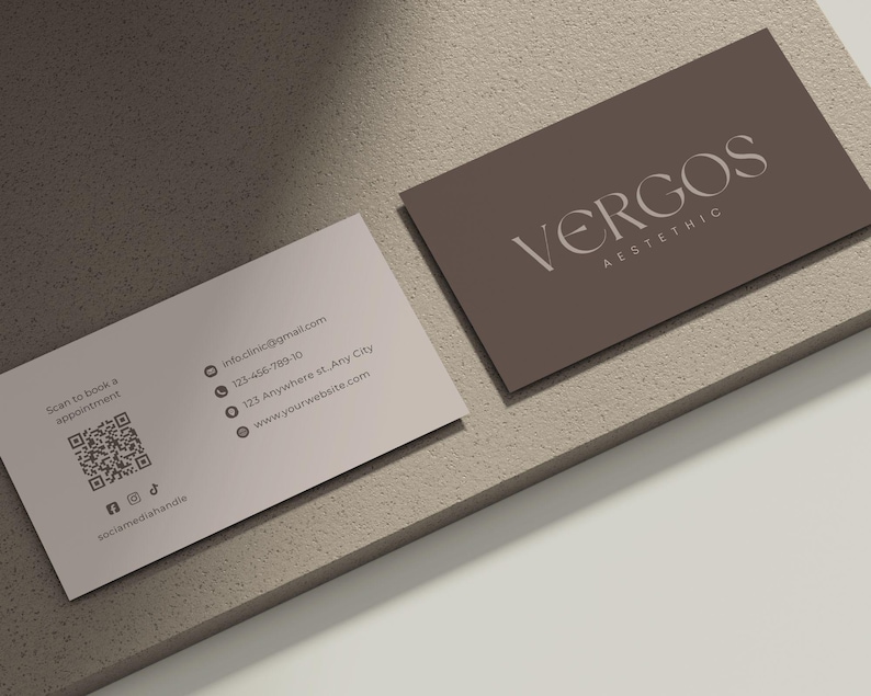 以下が含まれることがあります： 2 つの名刺。1 つは「VERGOS AESTHETIC」の白文字が入ったトープ色、もう 1 つは濃い灰色の文字と連絡先情報が入った薄いピンク色です。カードは、質感のある明るい茶色の表面にあります。