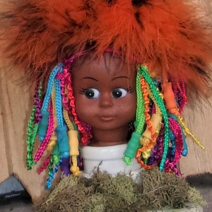 May include: A doll with a black face and brown eyes, wearing a bright orange fuzzy hat and colorful braids with beads. The doll is sitting on a base covered in green moss.